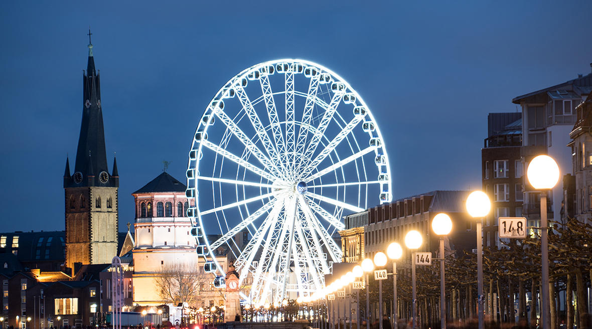 Illuminated ferris wheel next to two historic buildings with a row of bright street lamps