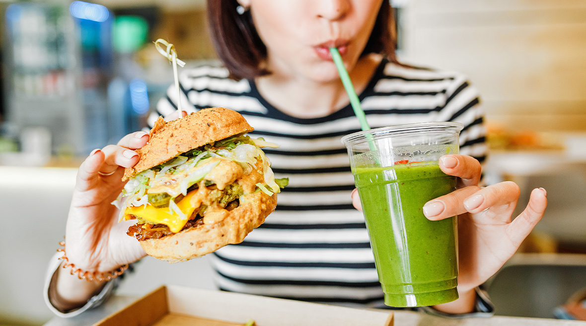 A young woman drinks a green smoothie from a straw while holding a large burger