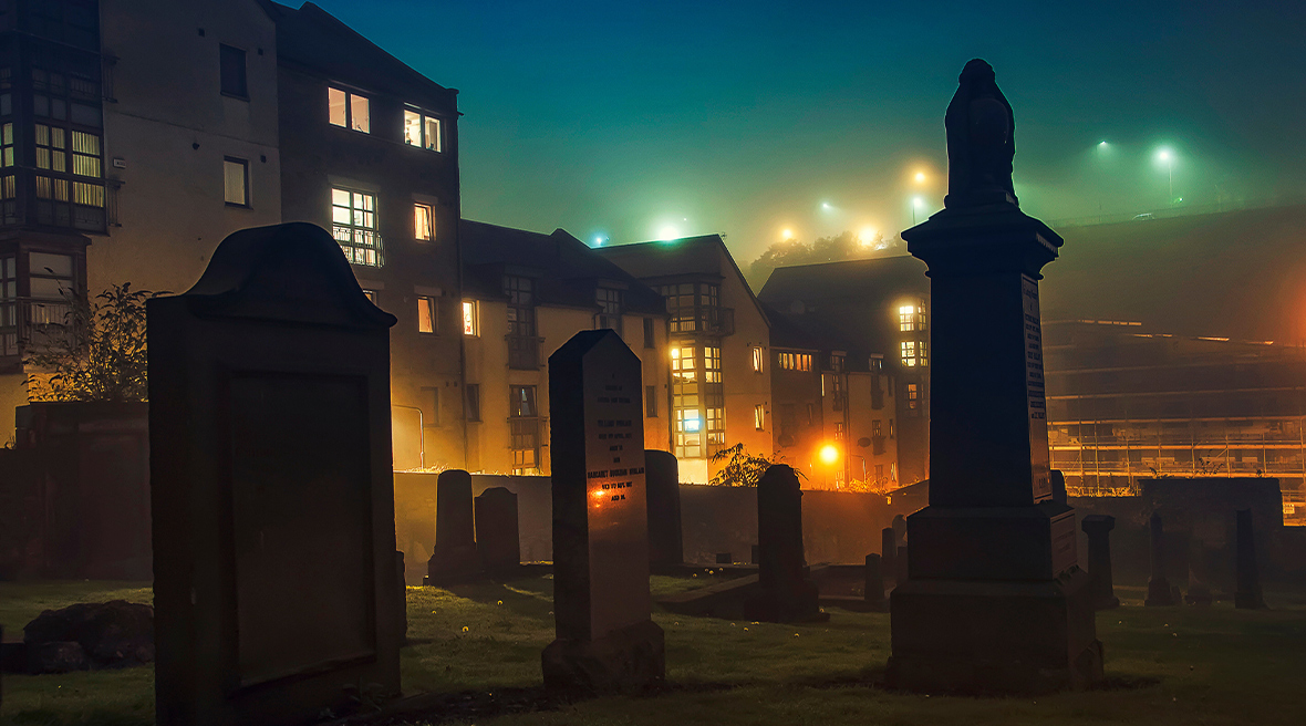Grafstenen bij nacht op een kerkhof voor huizen in een door straatlantaarns verlichte straat