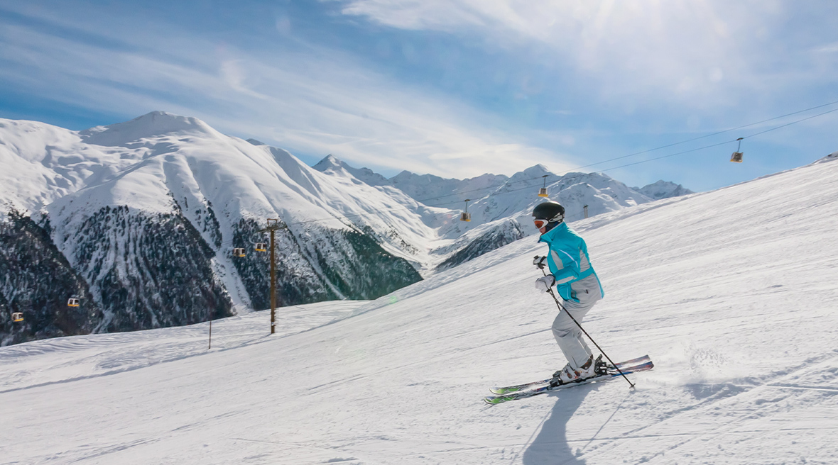 A skier in a light blue jacket skis down a slope of a ski resort, with a mountain range and a ski lift in the background