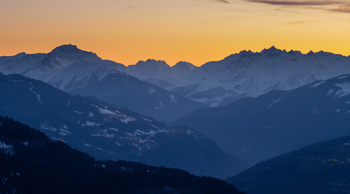 Orange sunrise over the mountains of La Rosière with lights of a town visible on the mountain side.