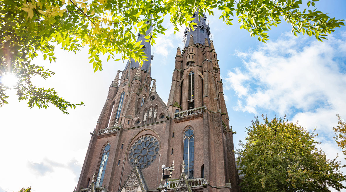 Two towers of a neo-Gothic or medieval church rising up into a blue sky with leafy trees framing the scene