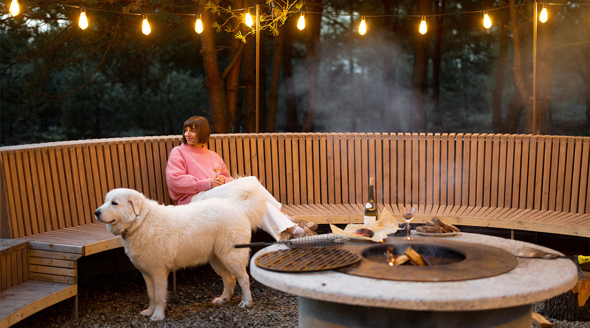 Young woman relaxing in an outdoor dining area with a dog, round a barbecue or fire with bottle of wine and glasses