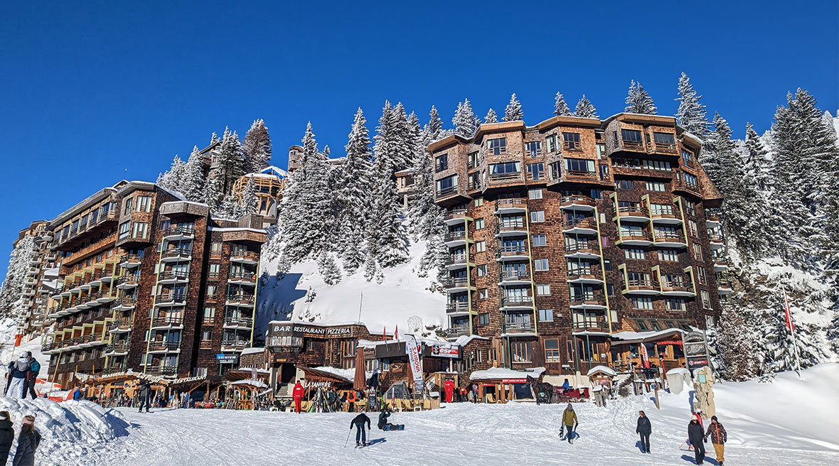 Wooden-fronted seven-storey apartments line up along the side of a piste with snowy fir trees and bright blue sky behind