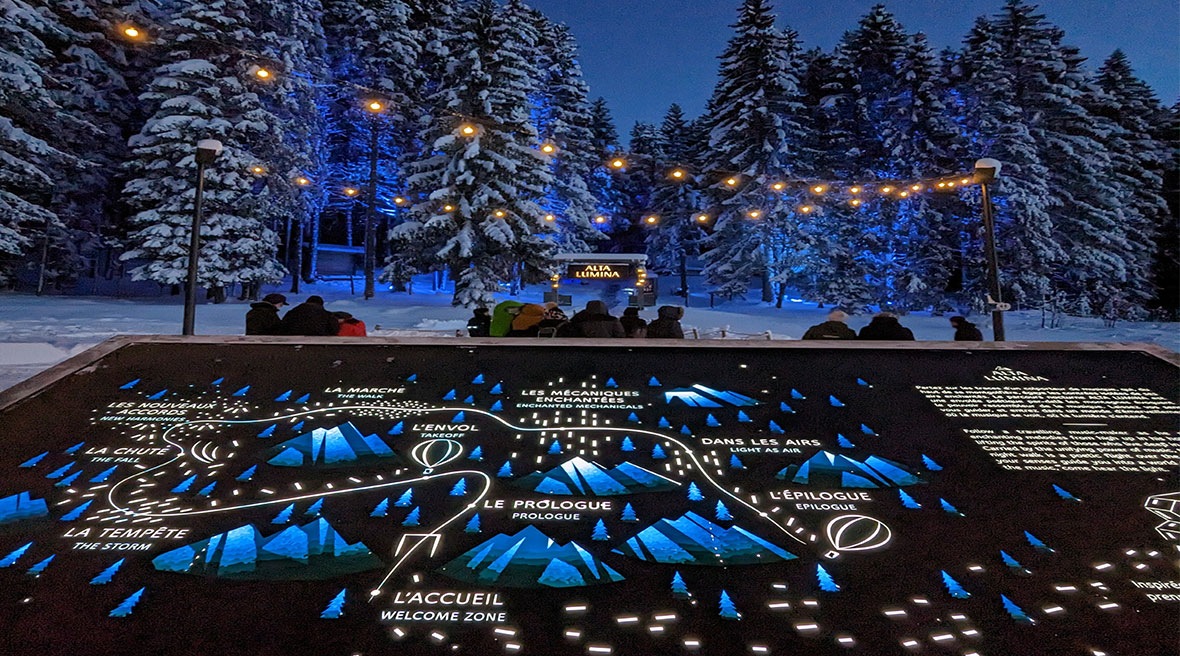 an illuminated information board in the foreground behind which lights lead the way to a clearing in the snowy trees and a bright sign saying Alta Lumina