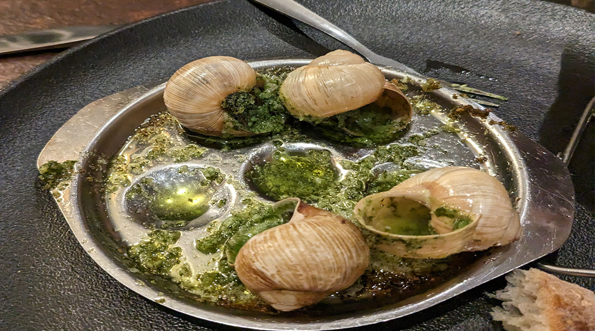 Four snail shells sit in buttery green juice on a metal snail dish. A torn piece of baguette sits next to the plate