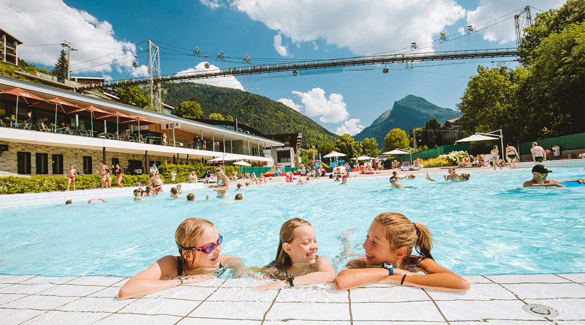 Three teenage girls chat in the water at the side the Morzine outdoor swimming pool, with others swimming behind them. In the background, the high up pedestrian bridge links both upper sides of the town against a sunny sky.