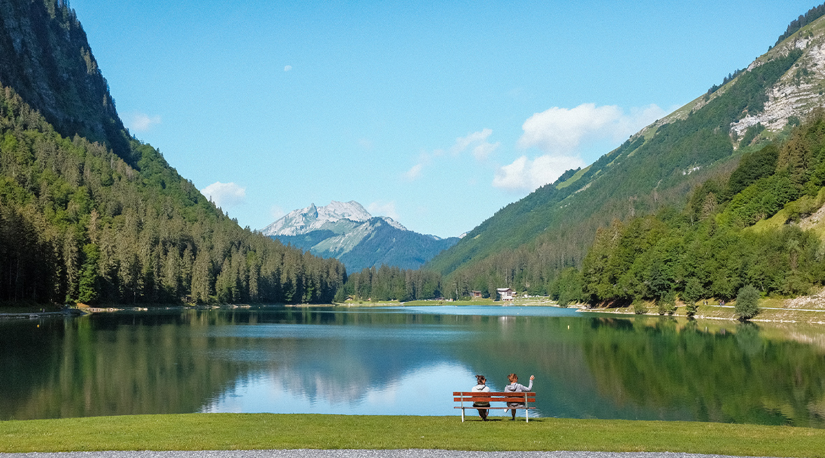 Two women sit on a bench in front of a large lake, gazing over the water. They are surrounded by green pine trees and we can see mountains in the distance, framed by a blue summer sky.