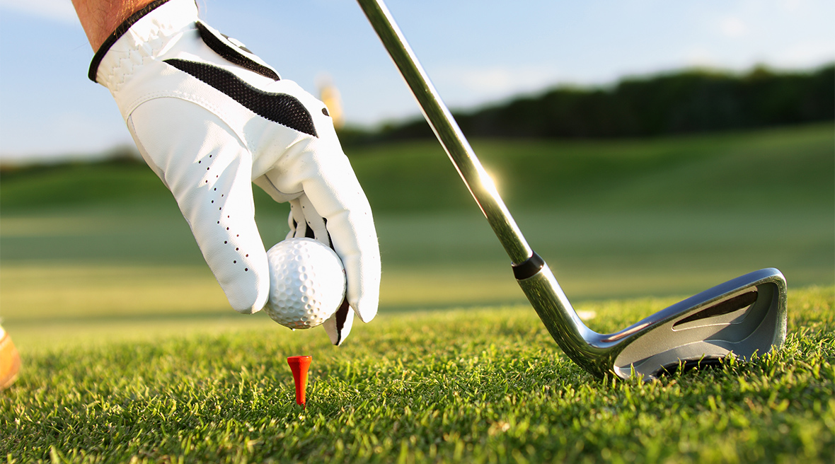 The hand of a golfer places a golf ball on a tee with an iron golf club on the right