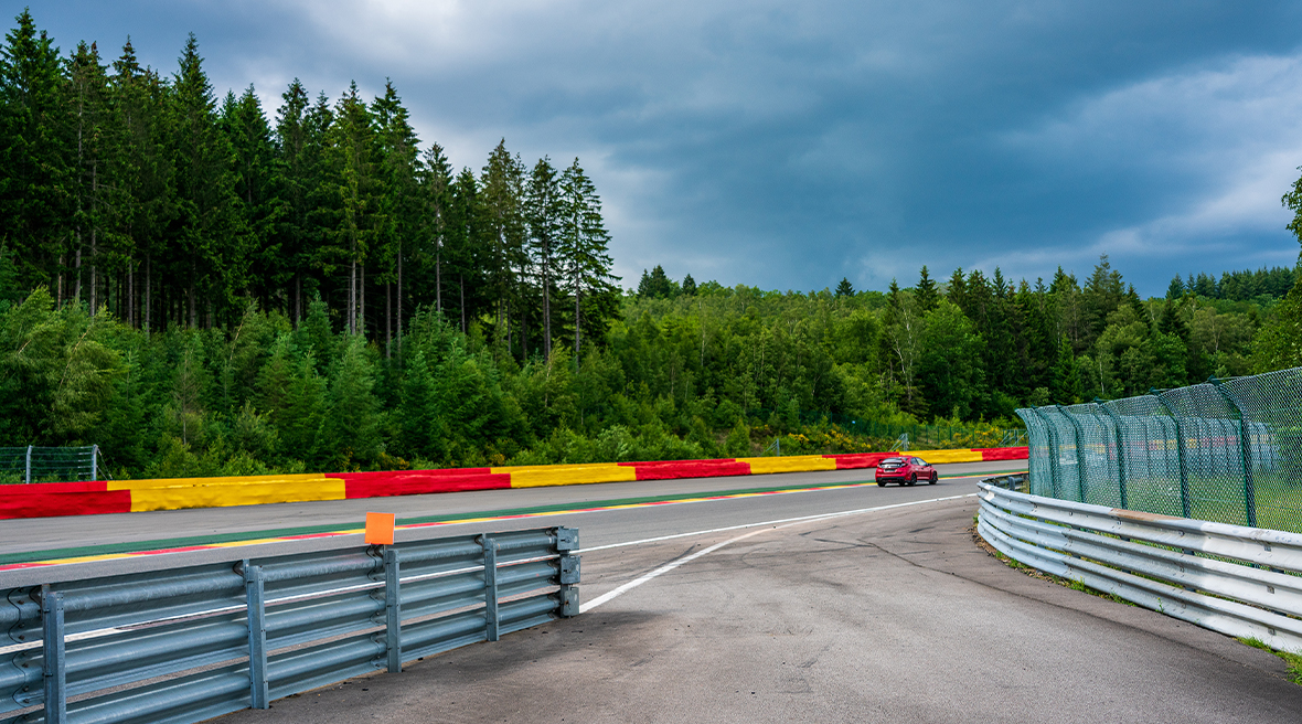 Red car driving round a bend on a motor racing circuit, with crash barriers either side of the track and forest outside the track.