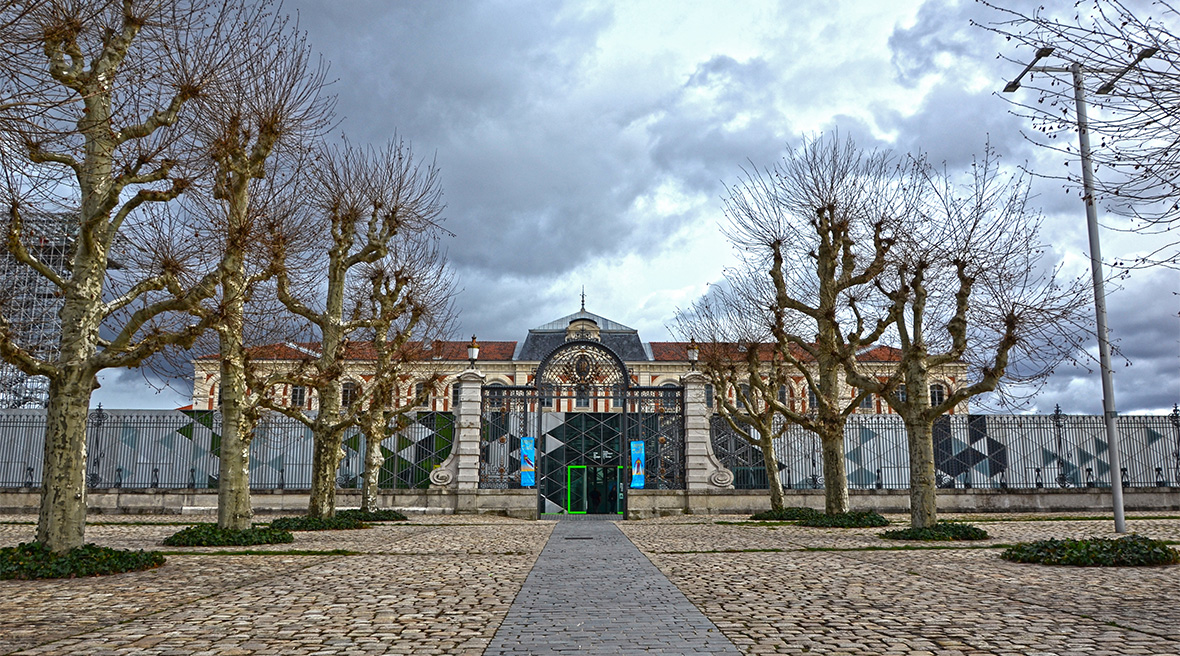 Leafless trees arranged symmetrically in front of a grand building and a lower one storey building, with an ornate black gate