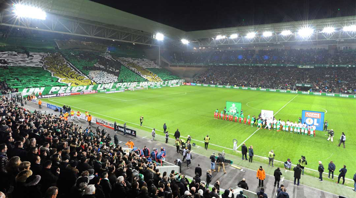 A football stadium under floodlights before a match. The stands are full of people and the teams are lining up on the pitch