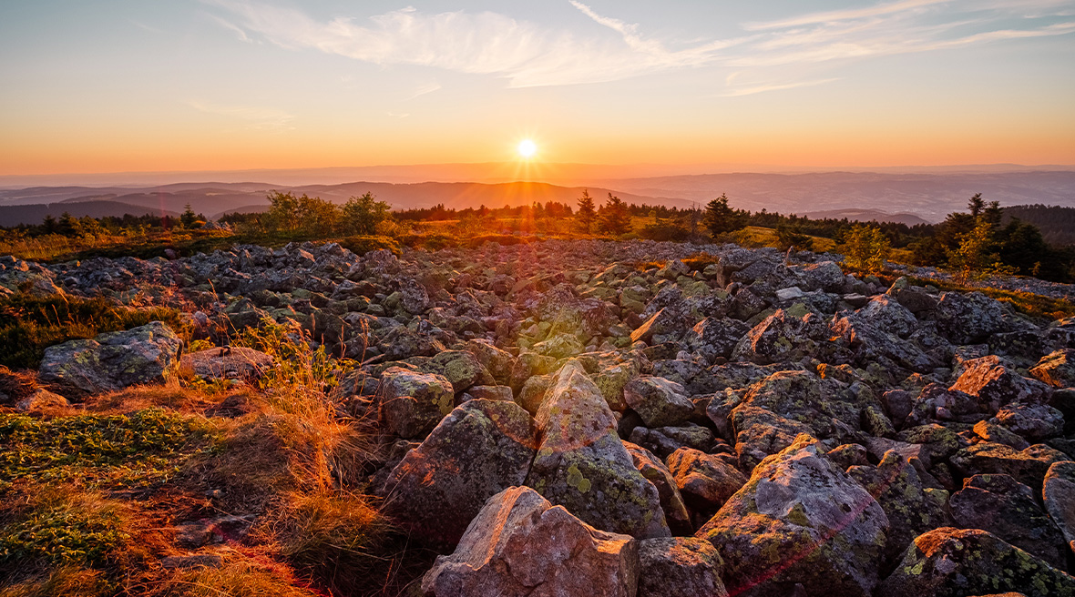 The sun rising over the horizon of a mountainous area, with large rocks in the foreground on a plateau