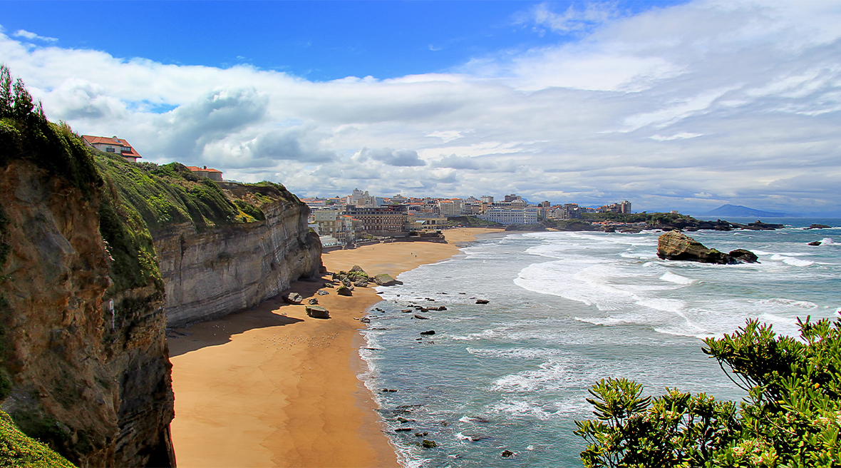 Towering cliffs, a long sandy beach with tides rolling in on a summer’s day, with blue sky, white clouds and a seaside town 