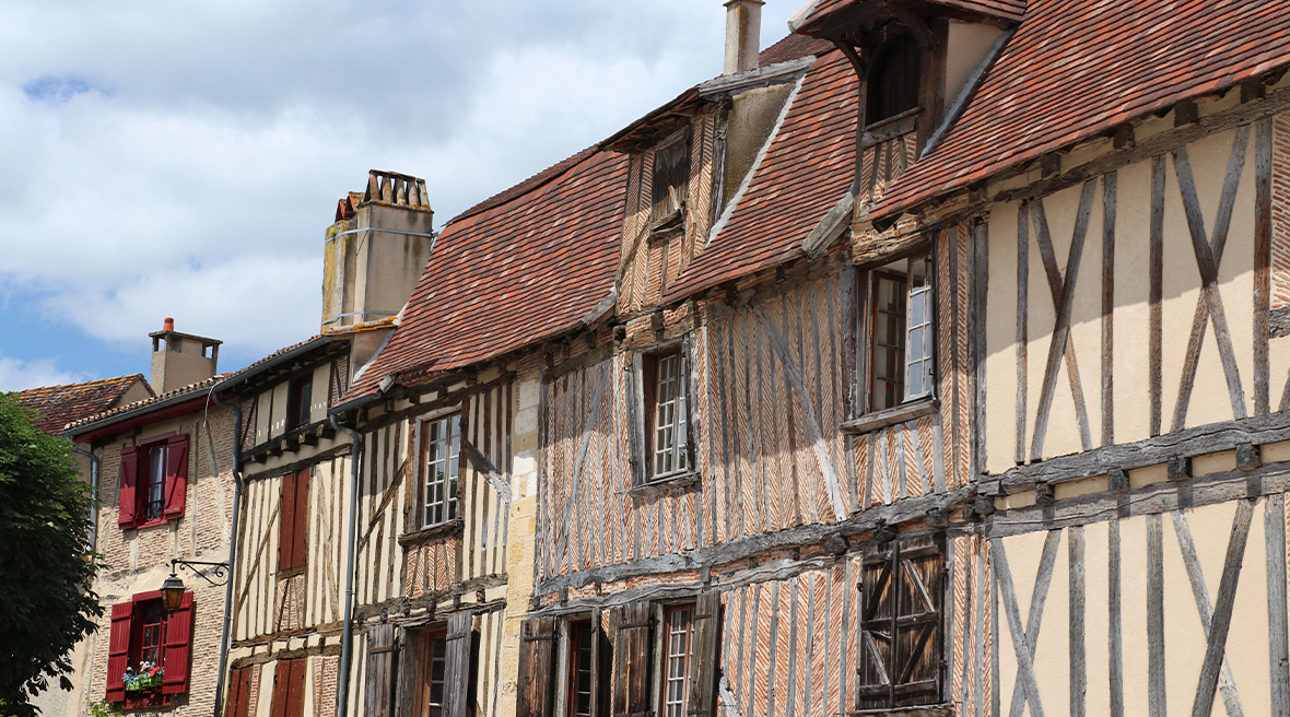 Five half-timbered old houses with coloured shuttered windows and chimneys with a cloudy blue sky above.