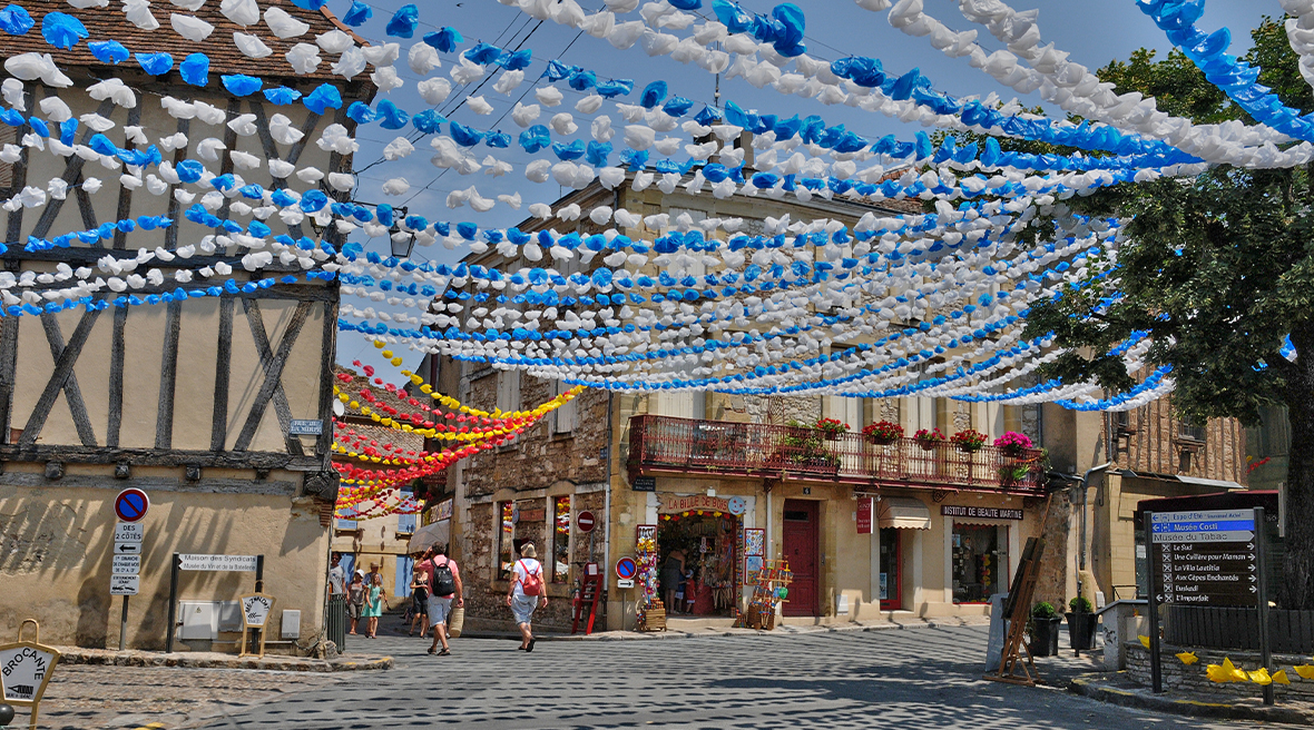 Colourful blue and white garlands hang between old buildings as people stroll down a pedestrianized street in Bergerac France.
