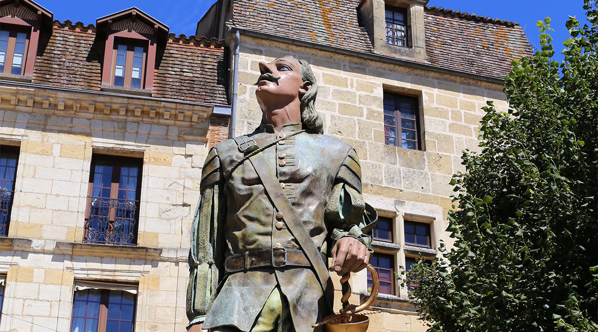Statue of soldier with large nose, holding his hat, looking upward in front of buildings, one of which is a ‘Foie Gras’ shop.