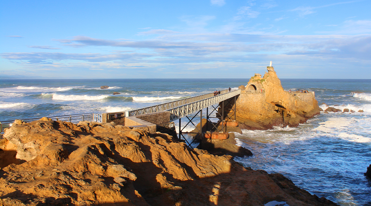 A bridge stretches between two rock formations in the sea, amid crashing waves