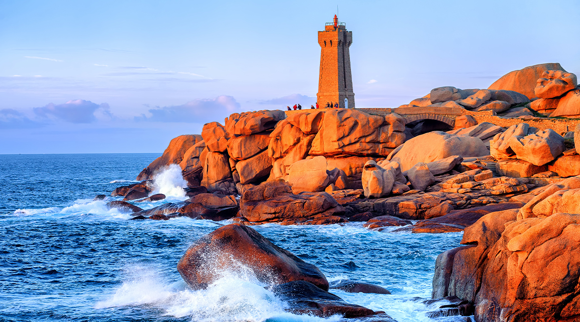 Rocky shore at sunset with lighthouse, waves crashing against the rocks.